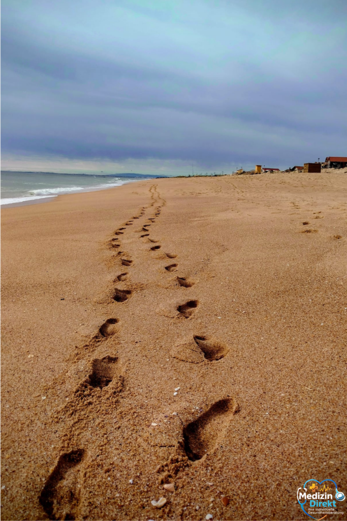 Fußspuren im Sand führen entlang eines ruhigen Strandes mit Meer und bewölktem Himmel.
